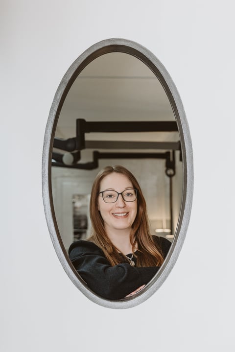 Woman with brown hair and glasses smiling in an oval mirror reflection against a white wall