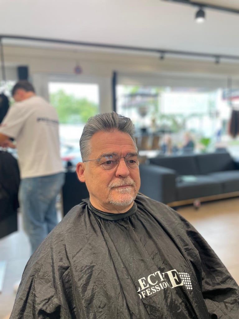 Middle-aged man with gray hair and glasses wearing a dark barber cape in a salon with other people visible in the background