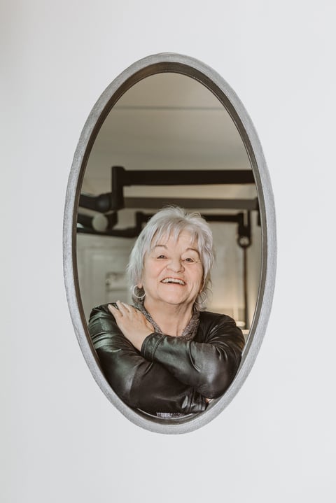 Portrait of an elderly woman with white hair wearing a black leather jacket, reflected in an oval mirror with a gray frame