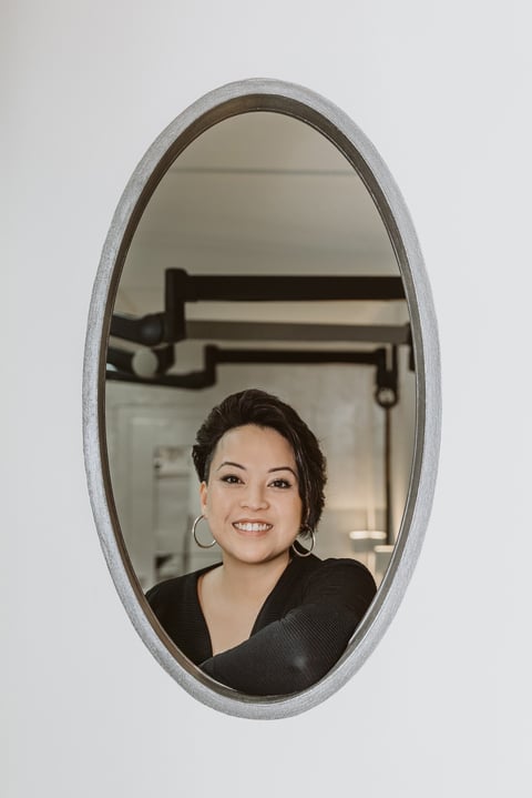 Woman with dark hair and hoop earrings smiling in an oval mirror mounted on white wall